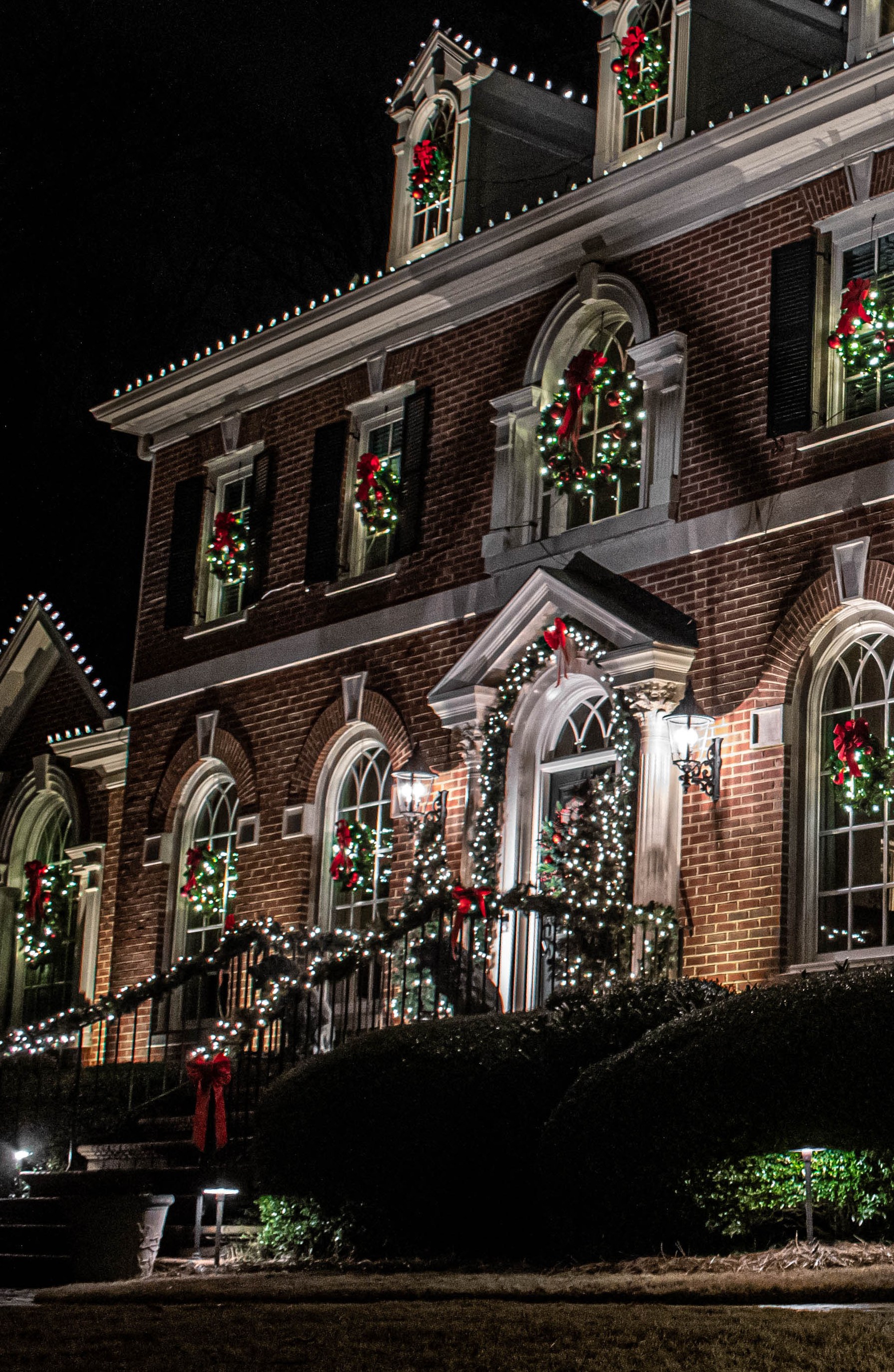 Brick home entry at night, lit wreaths with red ribbon and garland over the door